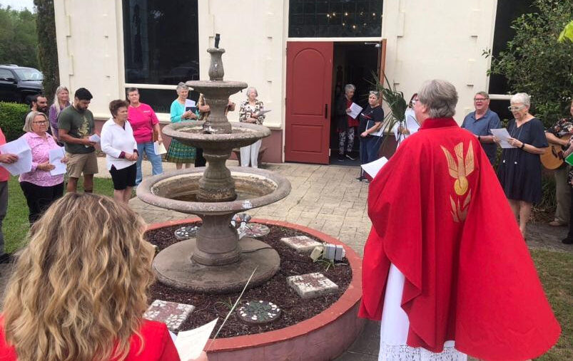 A priest in red vestments leading outdoor Pentecost service with a group of congregants singing outside the church in front of the red doors.